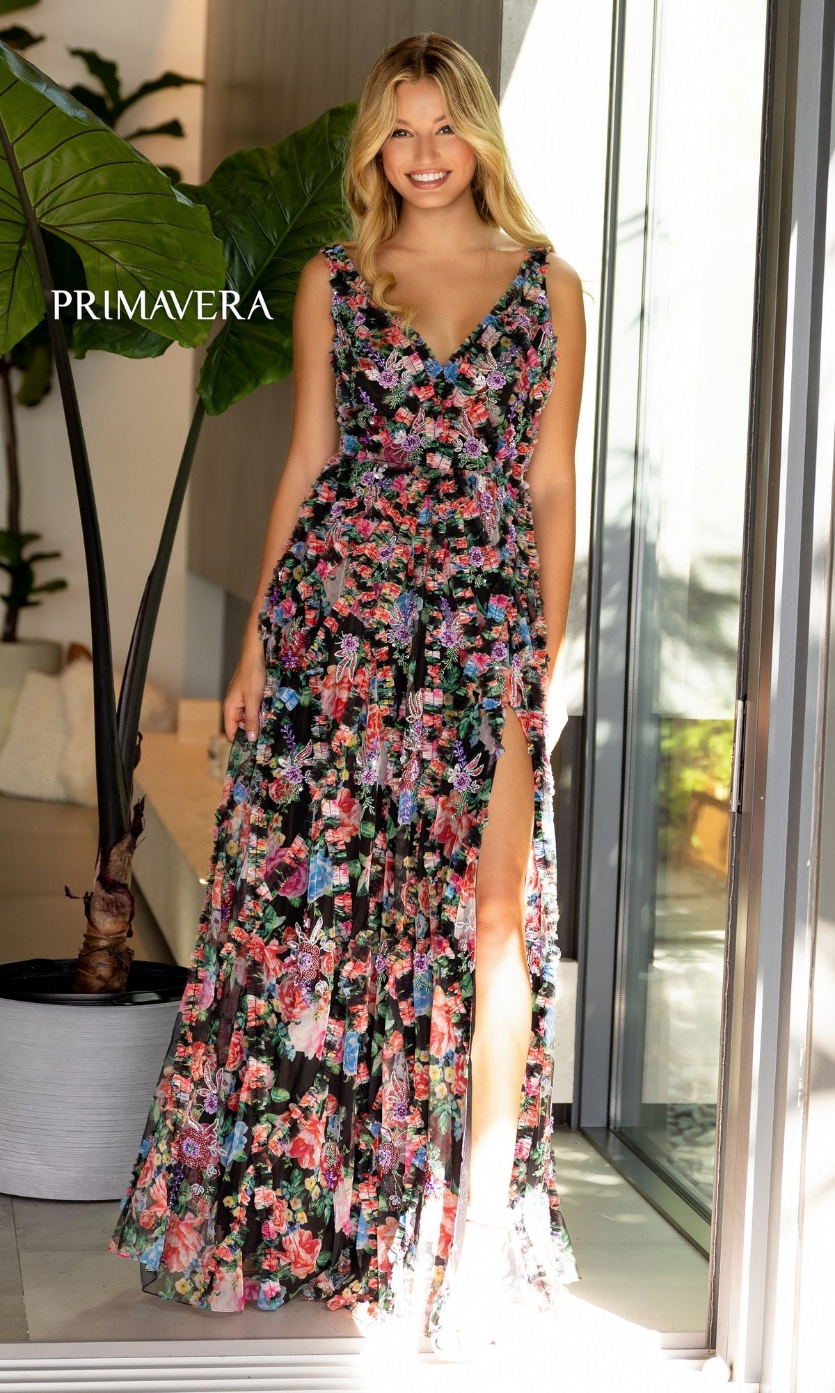 A woman stands indoors in the Primavera Long Print A-Line Ruffle Prom Dress 4167—a sleeveless floral prom gown with a thigh-high slit—smiling confidently with her hand on her hip.